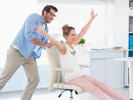 Ein Mitarbeiter schiebt eine mit ausgestreckten Armen und Beinen freudige Kollegin auf einem Bürostuhl durch den Raum